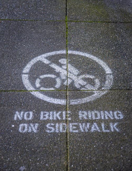Close-up view of a concrete sidewalk with a white painted circular symbol featuring a bicycle crossed out, indicating no bicycle riding permitted. Beneath the symbol, the text reads 'NO BICYCLE RIDING ON SIDEWALK'. The sidewalk consists of large rectangular slabs with visible expansion joints, and the surface shows slight texture and weathering. This image may be relevant to house removals or moving logistics when considering pedestrian and cyclist regulations in residential or urban areas, as seen in the context of parking and pavement rules for Mill Hill moves, as detailed on the page from Man with Van Mill Hill.