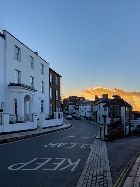 The image depicts a quiet residential street in Mill Hill during sunset, with a clear blue sky and orange clouds on the horizon. Prominent in the foreground is a white, multi-story house with modern architectural features, including a small balcony and large windows, situated on the left side of the image. The street curves gently to the right, with a steady flow of parked cars along the roadside. The roadway has clear markings, including the word 'KEEP' painted on the pavement, indicating parking restrictions. On the right side of the street, there is a pavement with tactile paving strips for pedestrian safety, along with black metal railings and a small flight of stairs leading up to a different level. The scene is well-lit by natural light, with no moving vehicles or pedestrians visible. This setting illustrates the typical environment where professional house removals and furniture transport services, such as those provided by Man with Van Mill Hill, operate during home relocation projects involving packing, loading, and transport of household items.