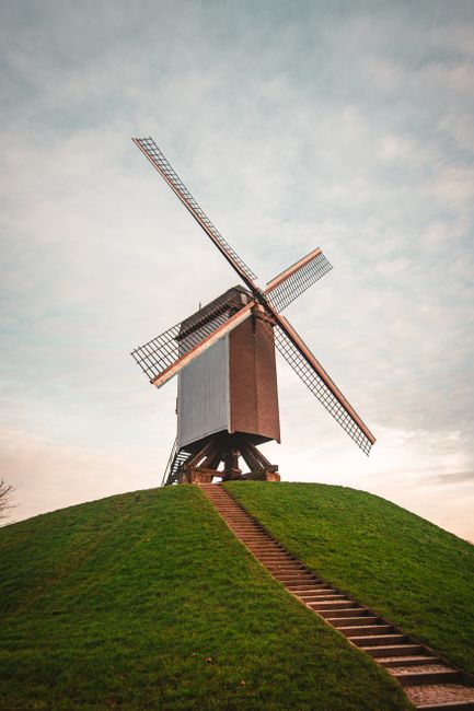 The image shows a traditional wooden windmill situated on top of a grassy hill, with a steep, straight set of stairs leading up to its entrance. The windmill has four large, lattice-style sails attach