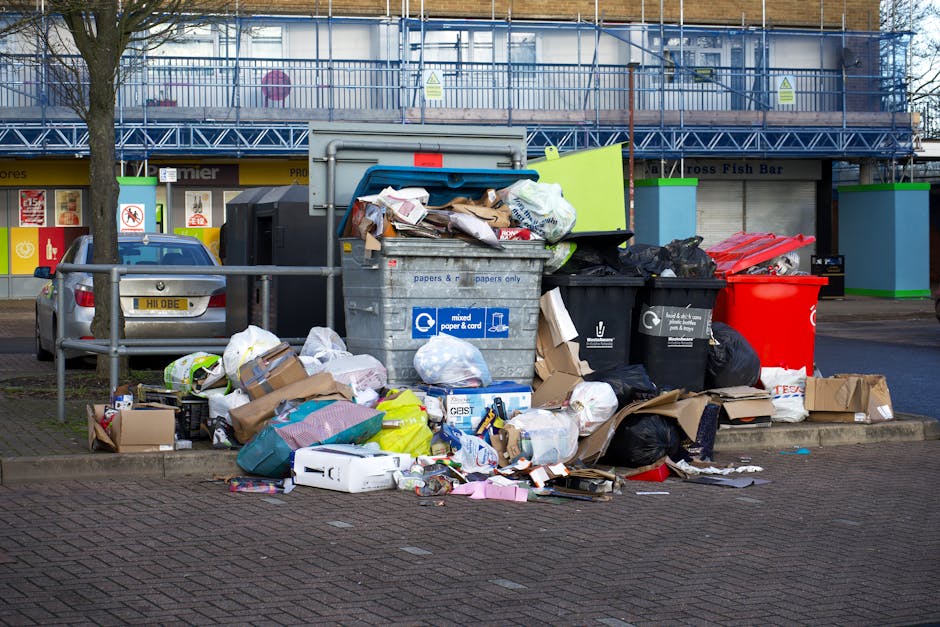 An outdoor scene in an urban setting showing a large pile of mixed waste and recyclables, including cardboard boxes, paper bags, black and red garbage bins, and plastic packaging, positioned on a paved area near a street. Several cardboard boxes and packing materials are scattered both next to and on top of the bins, with some items spilling onto the ground. Behind the waste collection area, there is a metal railing, two parked cars—one silver and one dark-colored—and a building with a glass façade under construction, covered with scaffolding and safety netting. The image captures a typical scene of waste clearance in a residential or commercial area, which is relevant to house and office removal services offered by Man with Van Mill Hill, highlighting the need for efficient bulk waste removal during home relocation or furniture transport processes.