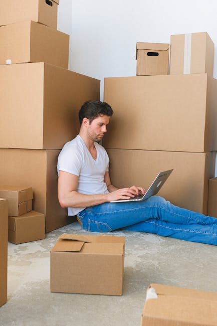 A young man with dark hair, dressed in a white t-shirt and blue jeans, is seated on the floor inside a room surrounded by numerous cardboard boxes of various sizes, some sealed with packing tape and o