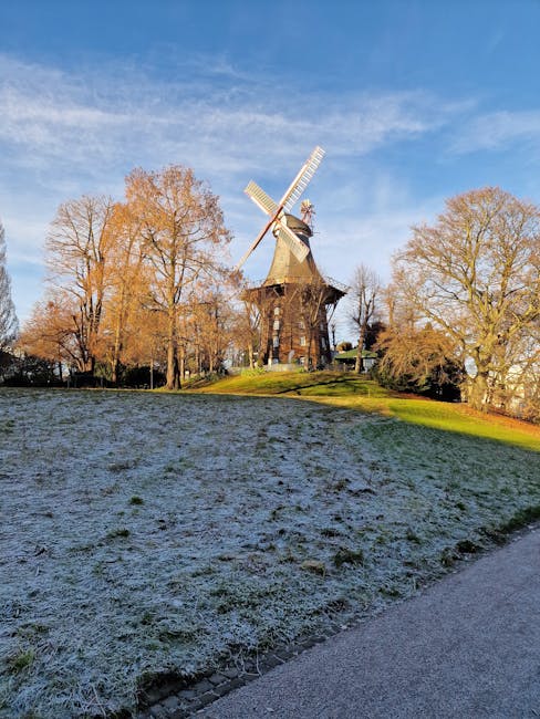 A tall wooden windmill with large white sails stands atop a grassy hill surrounded by leafless trees under a clear blue sky. The hill has a slight incline with patches of grass and a thin layer of frost, indicating cold weather. At the base of the hill, a paved pathway runs along the edge of the scene. The windmill's wooden structure is weathered, and the sails are positioned at an angle, suggesting a stationary posture. The setting appears to be a park or historic site near a residential or rural area, with the windmill serving as a prominent feature in the landscape. This image may relate to home relocation or moving services, highlighting the scenic environment around property sites managed by Man with Van Mill Hill during the process of packing and furniture transport.