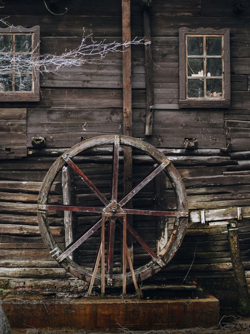 A large, weathered wooden water wheel mounted vertically on a rustic, dark-stained wooden exterior wall of a building. The wheel features prominent spokes and a circular frame, with some visible rust on the metal components. The building has two small, square windows with wooden frames, positioned above the water wheel. A bare tree branch extends into the upper left corner of the image, suggesting winter or early spring. The scene is outdoors, with the water wheel likely part of an old mill or rural structure. The overall setting emphasizes a vintage, rural aesthetic, relevant to home relocation or heritage property moving services offered by Man with Van Mill Hill, as captured during packing and loading processes involving historic or decorative exterior elements.