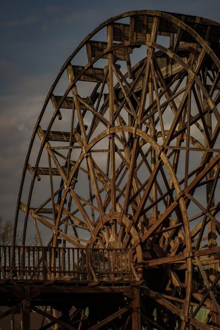 A large wooden Ferris wheel structure with a complex framework of interconnected wooden beams and supports, set outdoors against a cloudy sky. The wheel is partially visible, showing the intricate lattice of timber, with some sections appearing weathered. In the foreground, a wooden walkway or platform with railings surrounds the base of the Ferris wheel. This scene depicts an old amusement park or fairground ride, possibly during daylight hours with natural lighting. The image captures the scale and construction details of the vintage wooden ride, relevant to the context of heavy furniture and antique transport in home relocation or furniture transport services. Man with Van Mill Hill’s removals team might be involved in moving or restoring such large, delicate antique structures or similar sizeable items during house removals, where careful packing and moving logistics are essential.