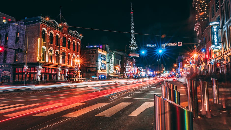 A night-time city street scene illuminated by vibrant neon signs and streetlights, with multiple commercial buildings on both sides featuring illuminated windows. Light trails from moving vehicles create streaks of red and white across the road, indicating long exposure photography. The street is equipped with metal bollards along the pavement to prevent vehicle encroachment, and the area appears busy with illuminated signs suggesting bars, shops, and entertainment venues. The environment is urban and lively, with visible overhead wires and a tall communication tower in the background. This scene is typical of a vibrant, bustling area suitable for a home relocation or furniture transport process, where careful planning by companies like Man with Van Mill Hill is essential for smooth moving and packing logistics.