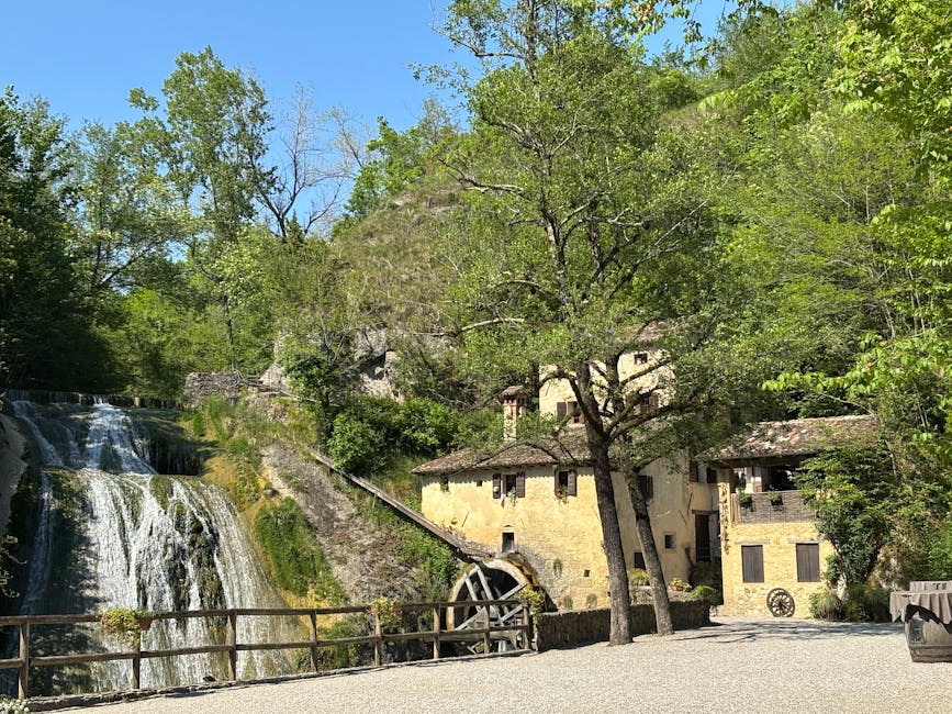 A scenic outdoor view featuring a historic stone building with small, dark windows and a tiled roof situated next to a cascading waterfall with multiple tiers. The waterfall flows over rocks into a small pond or stream, surrounded by lush green trees with bright foliage. In the foreground, there is a gravel pathway or open area with a wooden fence along the edge of the waterfall, and a large tree with a thick trunk and spreading branches partially obscures the building. The scene is well-lit by natural daylight under a clear blue sky, creating a tranquil setting that may be associated with residential or heritage sites, relevant to house removals or relocation services like those provided by Man with Van Mill Hill.