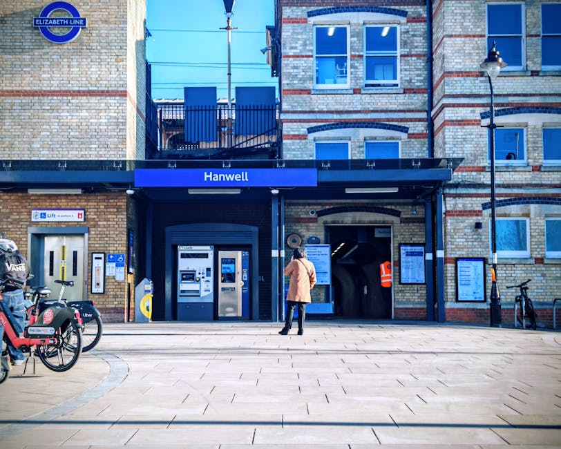 A woman wearing a beige coat, black trousers, and boots stands on the pavement outside the Hanwell station entrance, part of the Elizabeth Line, which features a brick facade with modern black accents and a blue sign displaying the station name. To her left, there are several bicycles, including a black bike with a delivery box labeled 'Vans' and another with a red basket, secured to a bike rack. The station entrance includes a ticket vending machine and a lift, with additional information boards mounted on the brick wall. The surrounding area is well-lit with street lamps, and a person wearing an orange high-visibility vest is visible near the staircase entrance to the station. The scene depicts a typical urban setting, relevant to house removals and furniture transport logistics, as detailed on the Man with Van Mill Hill service page about local removals near Mill Hill Broadway station.