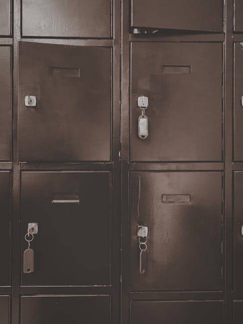 A close-up view of a set of dark metal storage lockers mounted on a wall, each with a rectangular slot for labels and a keyhole secured with padlocks. The lockers are arranged in a grid pattern, with visible hinges and handles on the front doors. Inside a property, a team from Man with Van Mill Hill is engaged in a home relocation process, evident from the presence of packing materials, cardboard boxes, and plastic-wrapped furniture in the background. The wooden table and various boxes are being carefully loaded or unloaded, utilizing equipment such as trolleys and straps to facilitate safe transport. The scene captures part of the furniture transport or packing and moving activities associated with professional removals services near NW7, highlighting the logistical aspects of a house move while emphasizing secure storage options for belongings during the process.