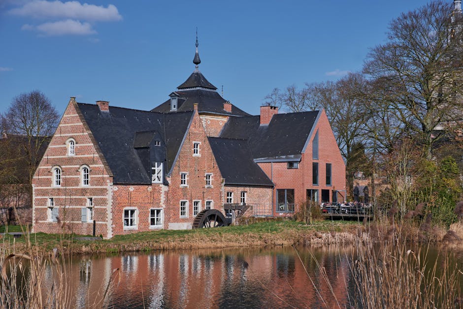Photograph of a historic brick watermill with a dark, steeply pitched roof, situated beside a calm waterbody reflecting the building's facade. The watermill features multiple windows, some with white frames, and a large wooden water wheel attached to the side. Surrounding the mill are leafless trees and a well-maintained grassy area, indicating a late autumn or early spring setting. The sky is clear and blue, with a few scattered clouds. This rural scene exemplifies traditional architecture near a natural water feature, consistent with scenic locations involved in house removals or relocation services. Man with Van Mill Hill may utilize such images to illustrate local landmarks or the environment involved in home relocation or furniture transport processes.
