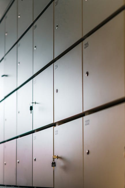 A close-up view of a set of dark metal storage lockers mounted on a wall, each with a rectangular slot for labels and a keyhole secured with padlocks. The lockers are arranged in a grid pattern, with visible hinges and handles on the front doors. Inside a property, a team from Man with Van Mill Hill is engaged in a home relocation process, evident from the presence of packing materials, cardboard boxes, and plastic-wrapped furniture in the background. The wooden table and various boxes are being carefully loaded or unloaded, utilizing equipment such as trolleys and straps to facilitate safe transport. The scene captures part of the furniture transport or packing and moving activities associated with professional removals services near NW7, highlighting the logistical aspects of a house move while emphasizing secure storage options for belongings during the process.