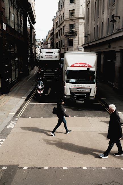 A street scene showing two white moving trucks parked adjacent to narrow multi-storey buildings in an urban area, with one truck labeled 'uniserve' on the cab and the other labeled 'Becton Transport' on its side. The trucks are positioned across a set of railway tracks embedded in the pavement, indicating a location with accessible loading points for house removals. Two pedestrians, a woman carrying a backpack and a man with white hair, are walking across a marked pedestrian crossing in the foreground, while a parked motorcycle is visible on the pavement near the trucks. The surrounding environment includes black external fire escapes and a mix of modern and older architectural facades, typical of a residential and commercial area. The scene captures a moment during home relocation logistics, with loading or unloading activities implied, and reflects the city’s urban moving process as managed by Man with Van Mill Hill.